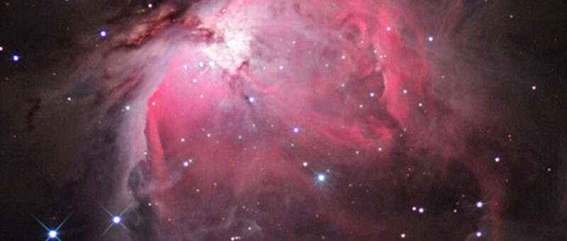 The Orion nebula, a hot pink cloud of gas with a bright white center and bright stars scattered around, is shown on a black background.