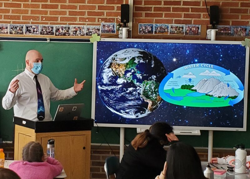 An astronomy educator guides students through the water cycle. The smartboard in the classroom displays an image of Earth and a graphic depicting the water cycle.