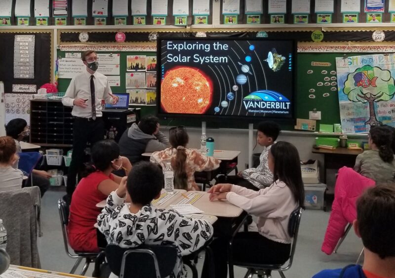 An astronomy educator talks to students in a classroom. On the smartboard, the title slide for "Exploring the Solar System" is displayed.