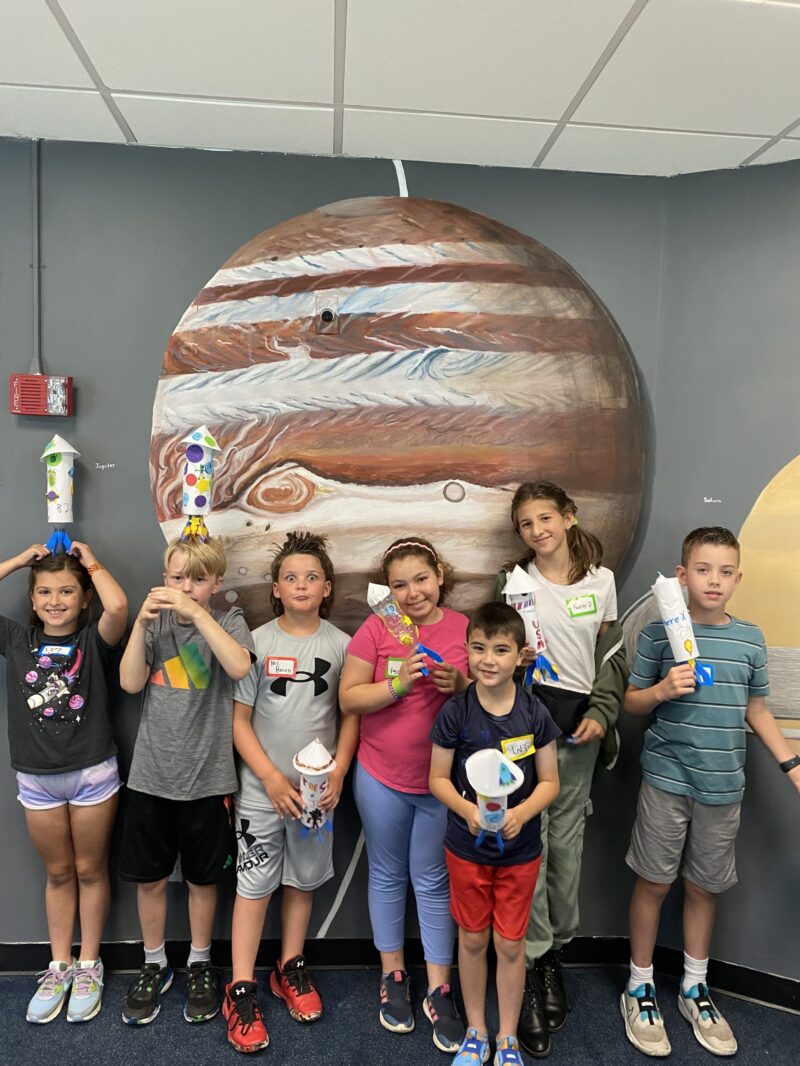 A group of Junior Astronomers stand in front of a mural of Jupiter.
